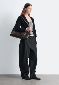 Schwarzer, strukturierter Cardigan über einem grauen Tanktop, kombiniert mit locker sitzenden schwarzen Hosen und flachen schwarzen Schuhen. Eine große, dunkle geflochtene Handtasche wird getragen.