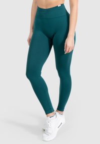 Teal hoge taille legging gemaakt van rekbaar materiaal met een gladde textuur, voorzien van discrete naden en gecombineerd met witte sneakers.