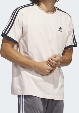 Osoba v světlobéžovém tričku Adidas s černými pruhy na rukávech a šedých kalhotách Adidas, ukazující ruce s prsteny a náramkem.