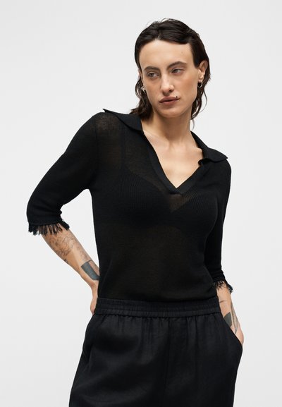 Μαύρο πλεκτό πουλόβερ με γιακά και V-λαιμόκοψη, μανίκια τριών τετάρτων και φθαρμένα άκρα μανικιών, φορεμένο με μαύρο παντελόνι με ελαστική μέση.