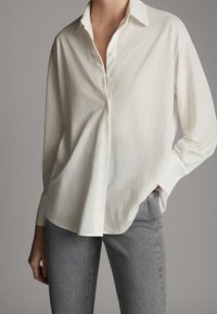 Vit långärmad skjorta med knappar har en avslappnad passform, krage, subtil textur och rundad nederkant, tillsammans med grå jeans.