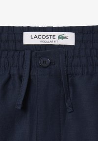 Pantalones cortos de corte regular en azul marino con cierre de botón y cintura elástica. Etiqueta de Lacoste mostrada en la parte superior con un logo de cocodrilo verde.
