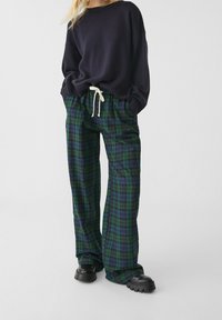 Czarny dzianinowy sweter z szerokimi rękawami, w połączeniu z zielono-niebieskimi spodniami w kratę z elastycznym pasem i sznurkiem, noszony z czarnymi butami.
