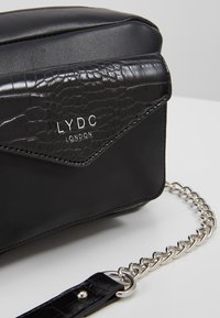 Borsa nera con un cappuccio effetto coccodrillo testurizzato. Presenta hardware in argento, una tracolla a catena e il logo impresso "LYDC LONDON". Tessuto del corpo liscio.