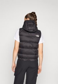 Zwarte gewatteerde vest met capuchon, voorzien van een gestikt ontwerp en logo-opdruk op de voorkant. Draag het over een wit shirt, gecombineerd met donkere broek.