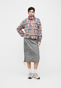 Άτομο στέκεται φορώντας πολύχρωμο πουλόβερ με σχέδια, γκρι midi cargo φούστα, λευκές κάλτσες και ανοιχτό ροζ αθλητικά παπούτσια σε απλό φόντο.