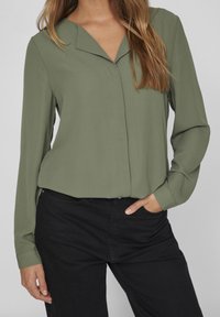 Blusa verde oliva con colletto, caratterizzata da una parte anteriore senza bottoni, maniche lunghe e un tessuto leggero e morbido. Abbinata a pantaloni neri.