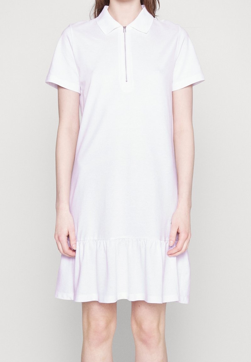 Femme portant une robe polo blanche à manches courtes avec fermeture éclair devant et ourlet à volants, debout devant un fond clair uni.