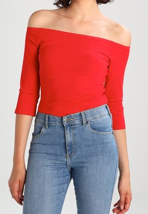Femme portant un haut rouge épaules dénudées avec des manches trois-quarts et un jean taille haute bleu clair, debout devant un fond uni.