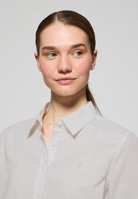 Biała koszula zapinana na guziki wykonana z lekkiego materiału o subtelnej fakturze. Posiada klasyczny kołnierzyk i małe haftowane logo na lewej piersi.