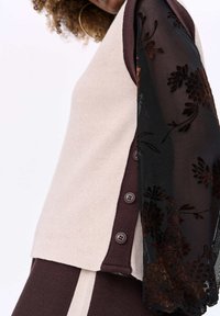 Sweter z beżowym korpusem, ciemnoczerwonymi akcentami i czarnymi przezroczystymi rękawami z haftem kwiatowym. Po jednej stronie znajdują się pięć ciemnych guzików.