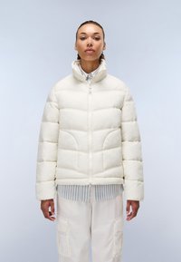 Hvid pufferjakke med lynlås foran, høj krave og quilted design. Båret over en blå og hvid stribet skjorte, der har sidelommer.