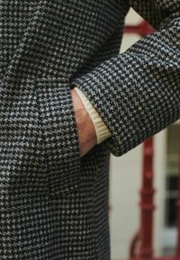 Caban negru și alb în carouri houndstooth, cu un material texturat, având un buzunar lateral și un mansetă de culoare deschisă care iese din manșetă.
