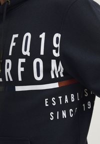Tengerészkék kapucnis pulóver, pamutból készült, nagy fehér szöveggrafikákkal, mint például "FQ19" és "ALAPÍTVA: 1987", színes kiegészítőkkel.
