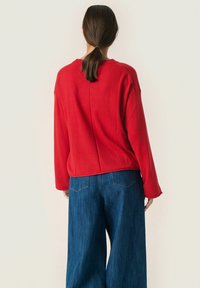 Rød, langærmet sweatshirt med rund halsudskæring og en afslappet pasform, parret med brede blå jeans, der viser en glat tekstur og en afslappet stil.