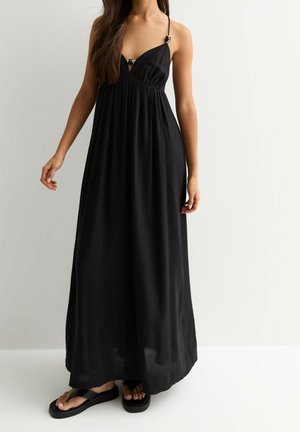 Schwarzes Maxikleid mit dünnen Trägern, V-Ausschnitt und geraffter Taille. Glatter Stoff mit fließendem Design. Kombiniert mit schwarzen Flip-Flops.