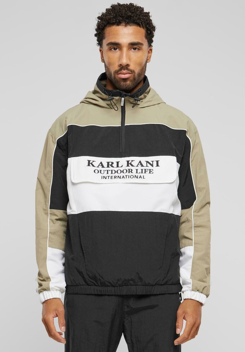 Vetrovka s farebnými blokmi v olivovej, čiernej a bielej farbe. Obsahuje zips na krku, elastické manžety a nápis "KARL KANI OUTDOOR LIFE INTERNATIONAL."