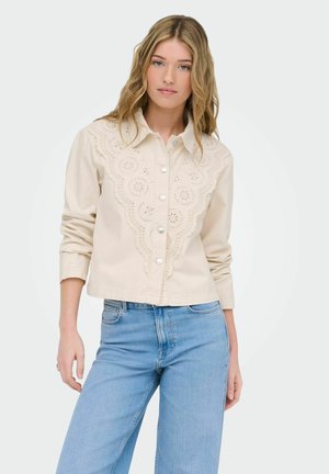 Giovane donna che indossa una camicetta color crema con bottoni e dettagli in pizzo a occhielli e jeans a vita alta di colore azzurro chiaro, in piedi davanti a uno sfondo semplice.