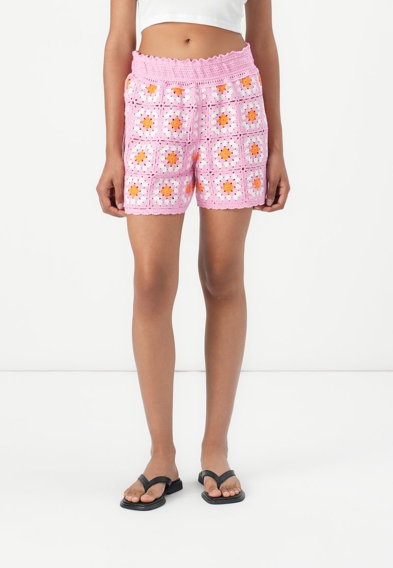 Femme portant un short en crochet rose avec des motifs floraux orange et blanc, un top court blanc et des tongs noires, debout sur un sol blanc.