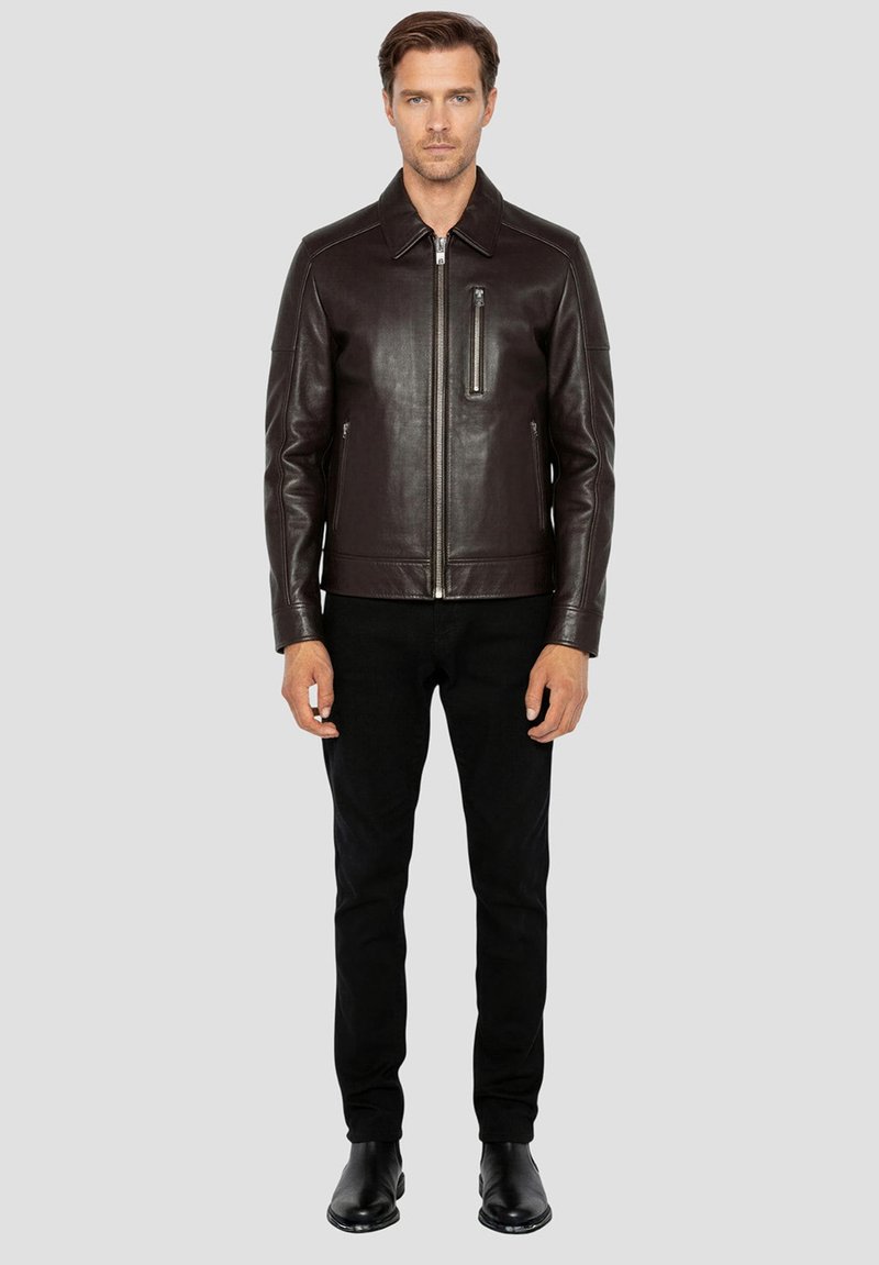 Bruine leren jas met een voorzijde rits, opstaande kraag, zijzakken en een met rits afsluitbare borstzak. Draag dit met een zwarte broek en laarzen.