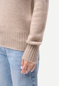 Manche de pull beige clair tricotée avec un poignet côtelé sur un jean denim bleu clair, main détendue sur le côté avec des ongles rose pâle.