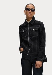 Svart denimjacka med dragkedja, två bröstfickor, kontrasterande vit sömnad och slagkrage. Bärs med mörka jeans och solglasögon.