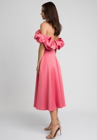 Różowa sukienka bez ramiączek z falbaną przy dekolcie, dopasowanym staniku i rozkloszowanym, midi dołem. Gładki materiał o delikatnym połysku. Eleganckie wysokie obcasy.