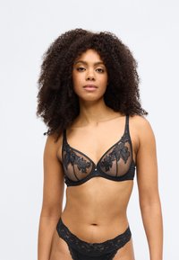 Μαύρο δαντελένιο σουτιέν με διαφανείς λεπτομέρειες, λουλουδάτα μοτίβα, στιβαρή κατασκευή και ρυθμιζόμενους ιμάντες. Διακοσμημένο με ένα μικρό διακοσμητικό φιόγκο στο κέντρο.