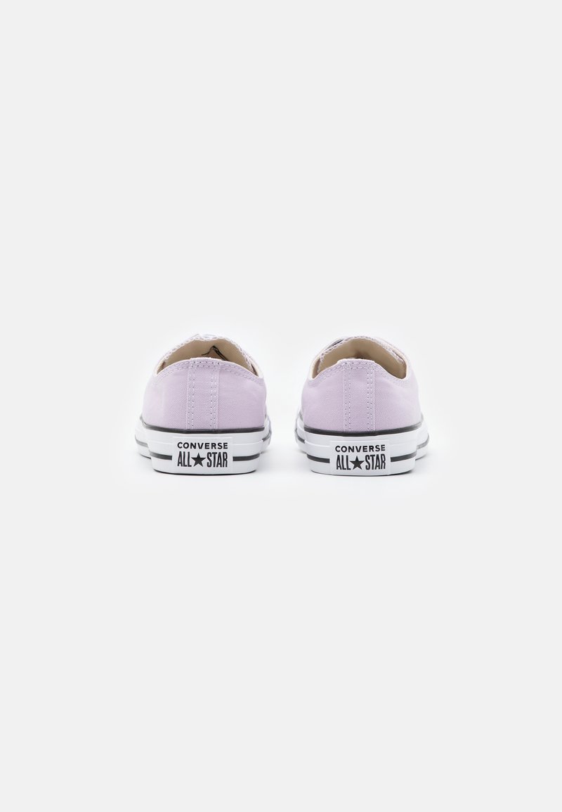 Zapatillas de lona lavanda de perfil bajo con cordones blancos, suela de goma negra y logo "Converse All Star" en el talón.