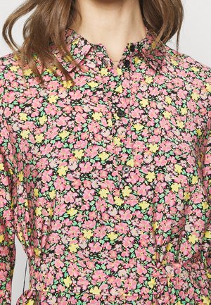 Kobieta ubrana w sukienkę w kwiaty z kołnierzykiem, z różowymi, żółtymi i zielonymi kwiatami oraz czarnymi guzikami z przodu.