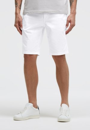 Mann trägt schlichte weiße knielange Shorts, weiße Turnschuhe mit schwarzem Fersendetail und ein in die Hose gestecktes weißes T-Shirt, steht vor einem weißen Hintergrund.