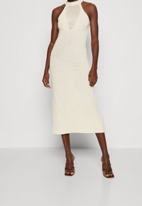 Robe tricotée couleur crème avec un col halter, décolletée à l'avant, longueur mi-mollet, présentant une texture à motifs et une silhouette ajustée.