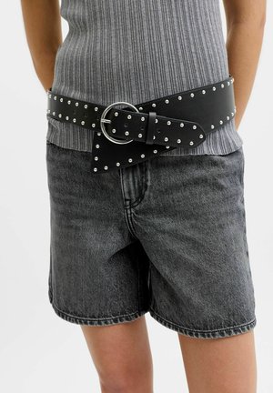 Osoba nosi crni kožni remen s ukrasnim zakovicama i velikom okruglom kopčom preko sive rebraste majice bez rukava i tamnosivih traper kratkih hlača.