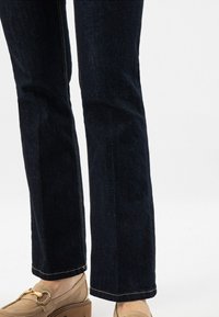 Dunkelblaue Jeans im geraden Schnitt, mit sichtbarer Naht, einem sauberen Saum und einer dezenten Textur, kombiniert mit beigen Slippers.