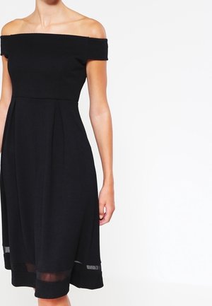 Kvinde iført en sort, knælængde kjole uden skuldre med gennemsigtig kantdetalje, stående foran en ensfarvet hvid baggrund.