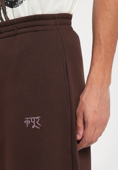Brown sweatpants made of soft fabric, featuring an elastic waistband and side pockets. Pink embroidered text detail on the thigh.
