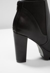 Czarny botek za kostkę z gładką skórzaną powierzchnią, zamkiem błyskawicznym, elastycznymi panelami oraz masywnym wysokim obcasem dla dodatkowej wysokości.