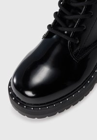 Stivaletto lucido nero con design lacciato, caratterizzato da una punta rotonda e una rifinitura borchiata lungo la suola. Texture liscia, suola in gomma robusta.