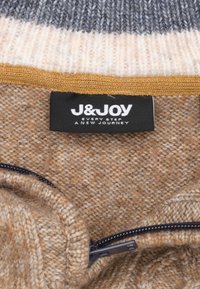 Pull en tricot beige avec un tissu texturé, présentant une fermeture éclair noire et une étiquette de marque. Le col arbore des rayures horizontales grises et crème.
