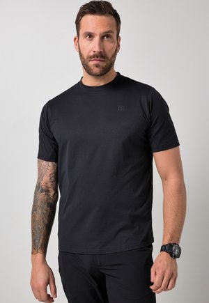 Mand med kort brunt hår og skæg iført sort t-shirt, sorte bukser, armbåndsur samt med en hel-ærmet tatovering på venstre arm, stående foran en ensfarvet baggrund.
