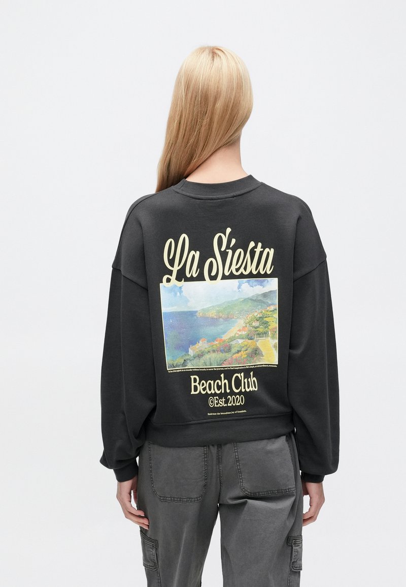 Μαύρο φούτερ με χαλαρή εφαρμογή, που διαθέτει πολύχρωμο γραφικό παραλίας και κείμενο "La Siesta Beach Club" στην πλάτη.