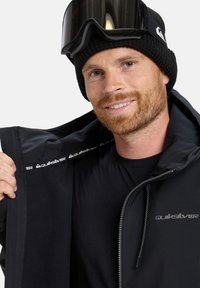 Black jacket with a smooth exterior and fleece lining, featuring Quiksilver branding inside, worn with a black knit beanie and goggles.