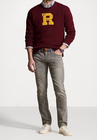 Burgunderfärgad stickad tröja med en gul "R"-logotyp, kombinerad med grå slim-fit jeans och beige sneakers. En blå och vit randig skjorta syns lite.