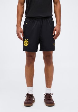 Hombre con pantalones cortos deportivos negros con el logo amarillo BVB 09, camiseta negra, calcetines blancos y zapatillas marrones, de pie frente a un fondo liso.