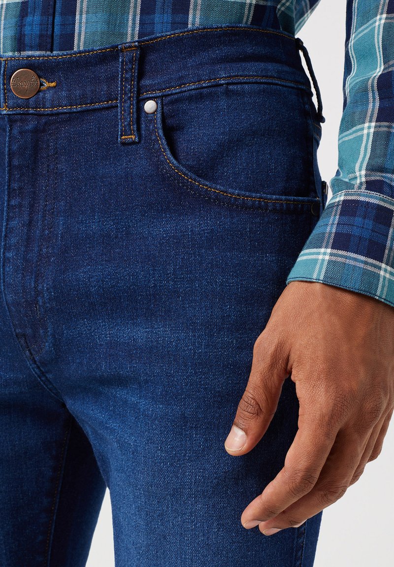 Ciemnoniebieskie dżinsy z bliskim ujęciem przedniej kieszeni i pasa, z pomarańczowymi przeszyciami i metalowym guzikiem.