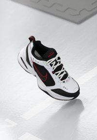 Adidașii albi Nike Air Monarch cu accente negre, logo roșu, panouri din plasă texturată și șireturi albe, expuși pe o suprafață reflectorizantă.