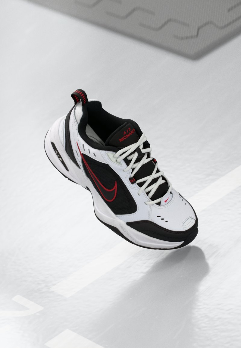 Adidașii albi Nike Air Monarch cu accente negre, logo roșu, panouri din plasă texturată și șireturi albe, expuși pe o suprafață reflectorizantă.