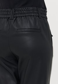 Pantalons en cuir noir avec une taille froncée, poches latérales et une texture lisse. Le design est ajusté sans fermetures visibles.