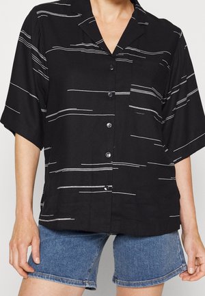 Czarna koszula na guziki z krótkim rękawem i białymi poziomymi paskami zestawiona ze średniej długości niebieskimi szortami jeansowymi na stojącej osobie.