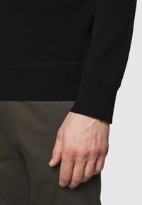 Maglione nero a maglia con polsini a coste, texture liscia e design lineare. Mano visibile contro pantaloni verde oliva con dettagli sottili.